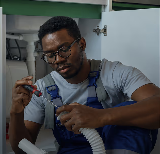 Man in glasses and overalls repairs plumbing under a sink using a screwdriver.